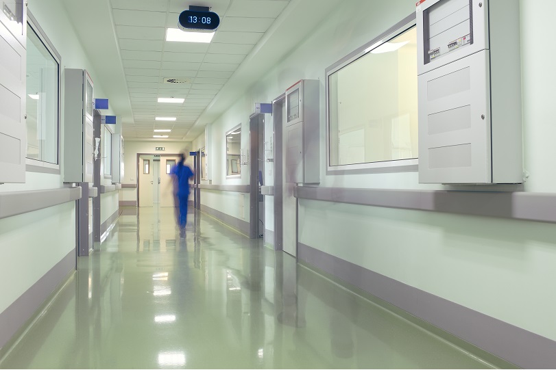 Foto ilustrativa mostra o corredor de hospital. Ao fundo, há a imagem desfocada de uma trabalhadora. Ela está de costas e usa uniforme azul. Predominam as cores branca e cinza.