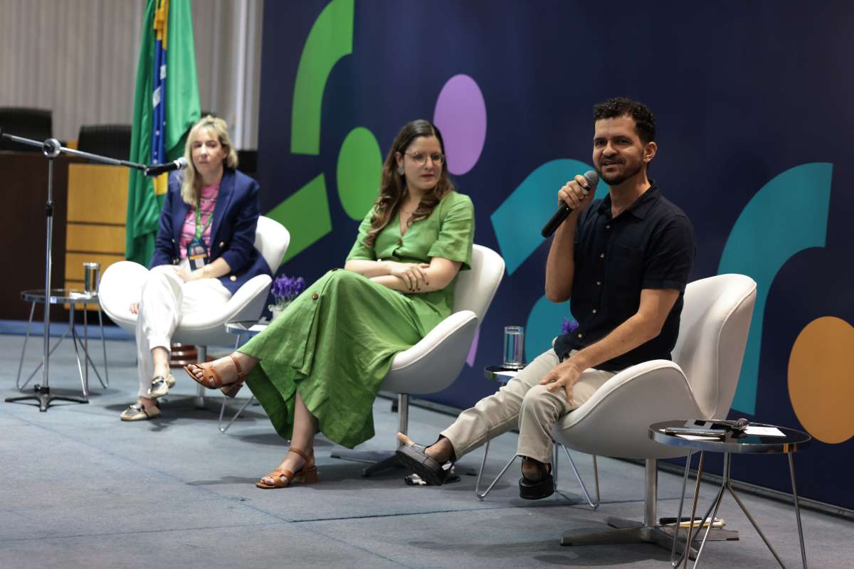 Foto de três pessoas sentadas, duas mulheres e um homem com microfone na mão discursando