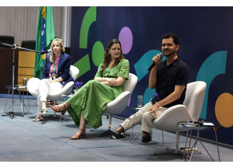 Foto de três pessoas sentadas, duas mulheres e um homem com microfone na mão discursando