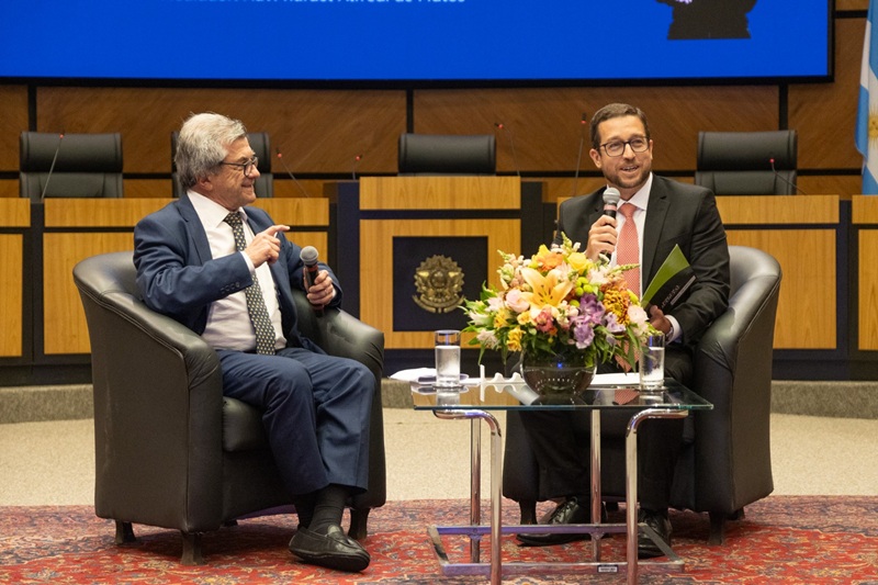 A fotografia mostra o palestrante Juan Delgue conversando com o mediador Rafael, ambos sentados em poltronas no plenário.