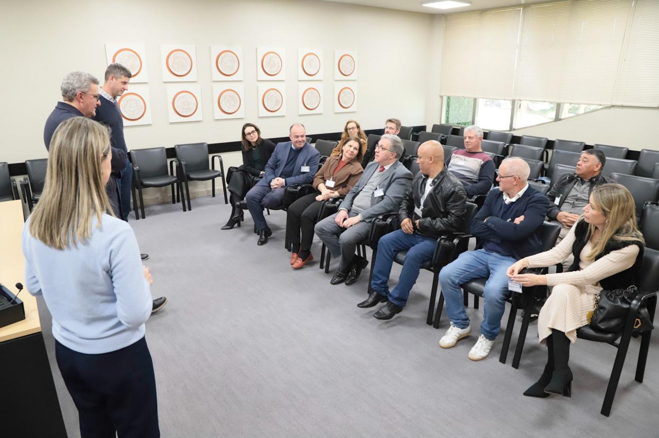 Três pessoas aparecem na esquerda de pé e na direita várias estão sentadas. 