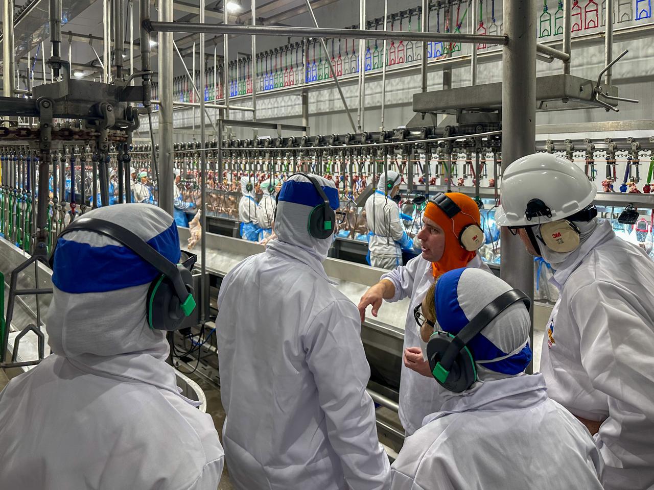 A foto mostra a comitiva do TRT visitando as instalações da linha de produção. As pessoas usam roupas de proteção brancas.