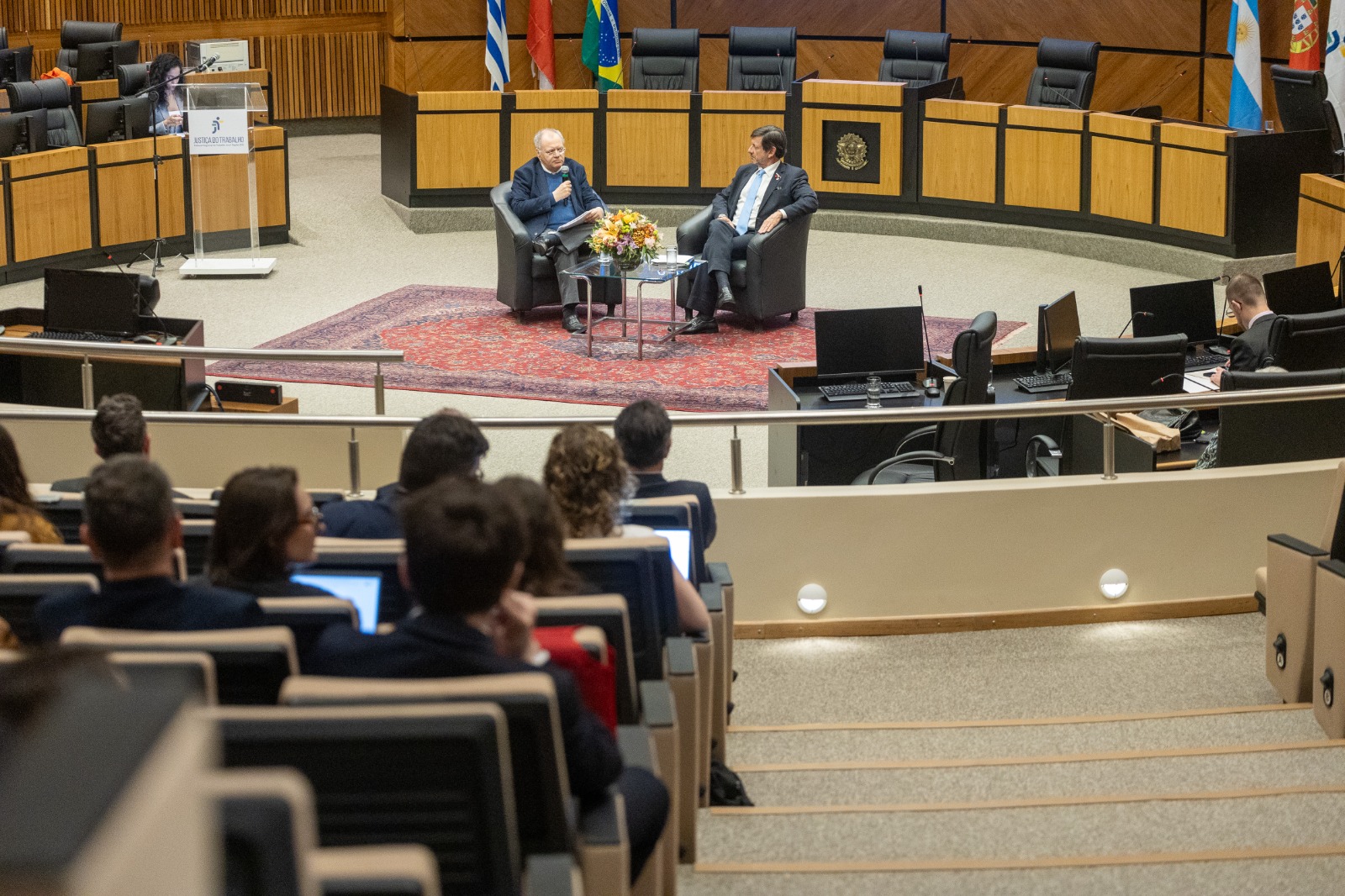 Foto em ângulo médio, mostrando parte do público nas poltronas e palestrantes na nave do plenário