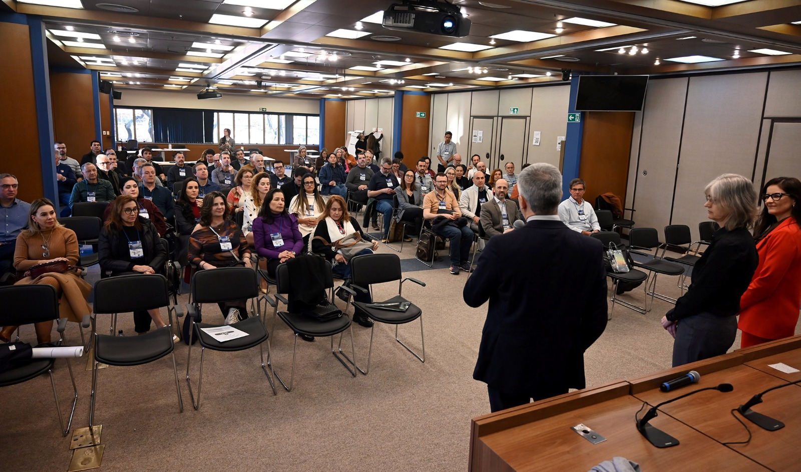 Foto em ângulo aberto, mostrando público sentado e palestrantes em pé