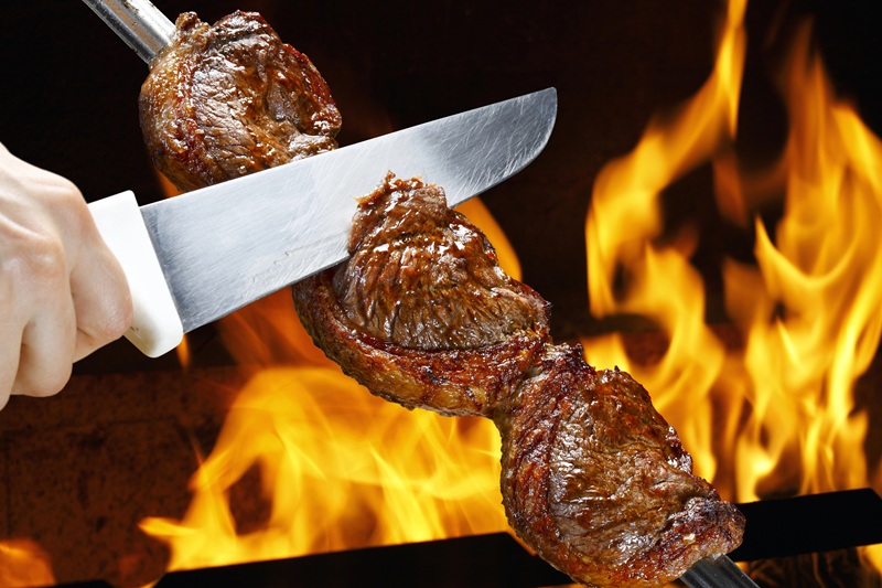 A foto mostra a mão de um assador, cortando uma peça de picanha com uma faca de churrasco. Atrás, há uma chama de fogo.