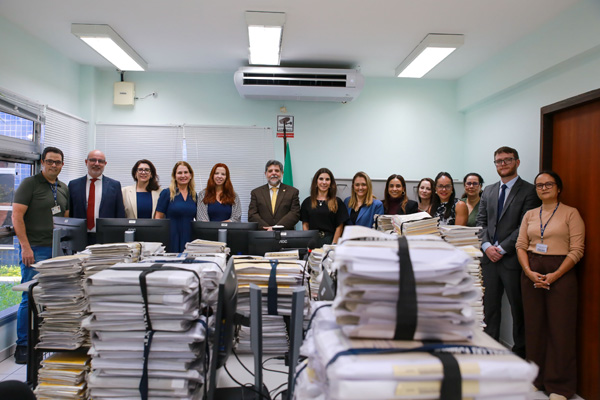 A imagem mostra várias pessoas lado a lado posando para foto com processos físicos na frente.