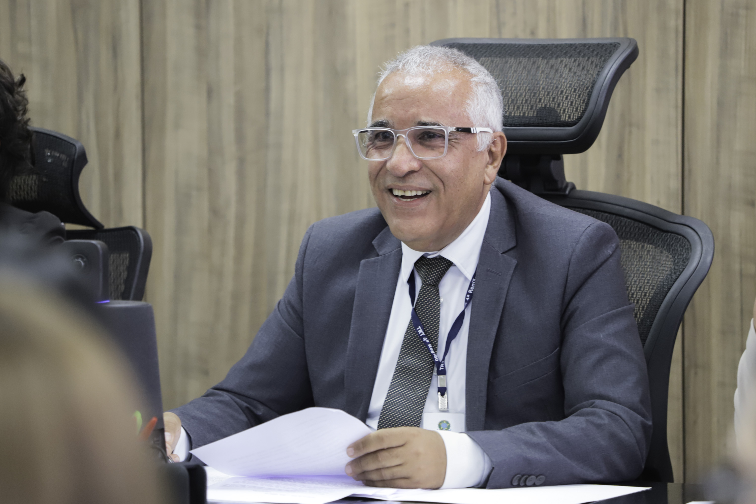 Gilberto é negro e grisalho. Usa terno cinza e óculos de armação grande. Está na mesa oficial, sorri olhando para a direita da imagem segura papéis.