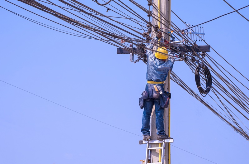 A fotografia mostra pessoa trabalhando em poste de luz, com equipamentos de proteção, e um céu azul ao fundo.
