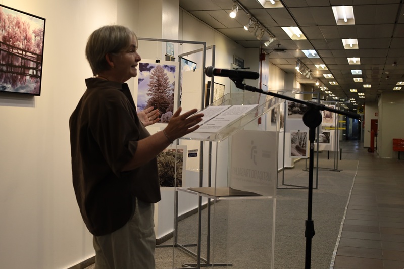 Imagem de Joice Kreiss discursando ao microfone no evento de abertura, ao fundo estão as fotos da exposição. Joice está de pé. Ela é uma mulher que aparenta ter em torno de 60 anos, tem cabelo curto branco, pele clara e veste uma camisa marrom e uma calça clara.