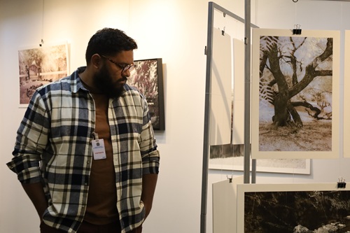 A imagem mostra um homem olhando as fotos da exposição. Ele está de pé, com a cabeça inclinada para baixo. Ele é um homem negro que aparenta ter 35 anos, de cabelos e barba escuros, veste uma camisa com estampada xadrez bege, com listras azuis aberta e por baixo dela uma camiseta marrom.