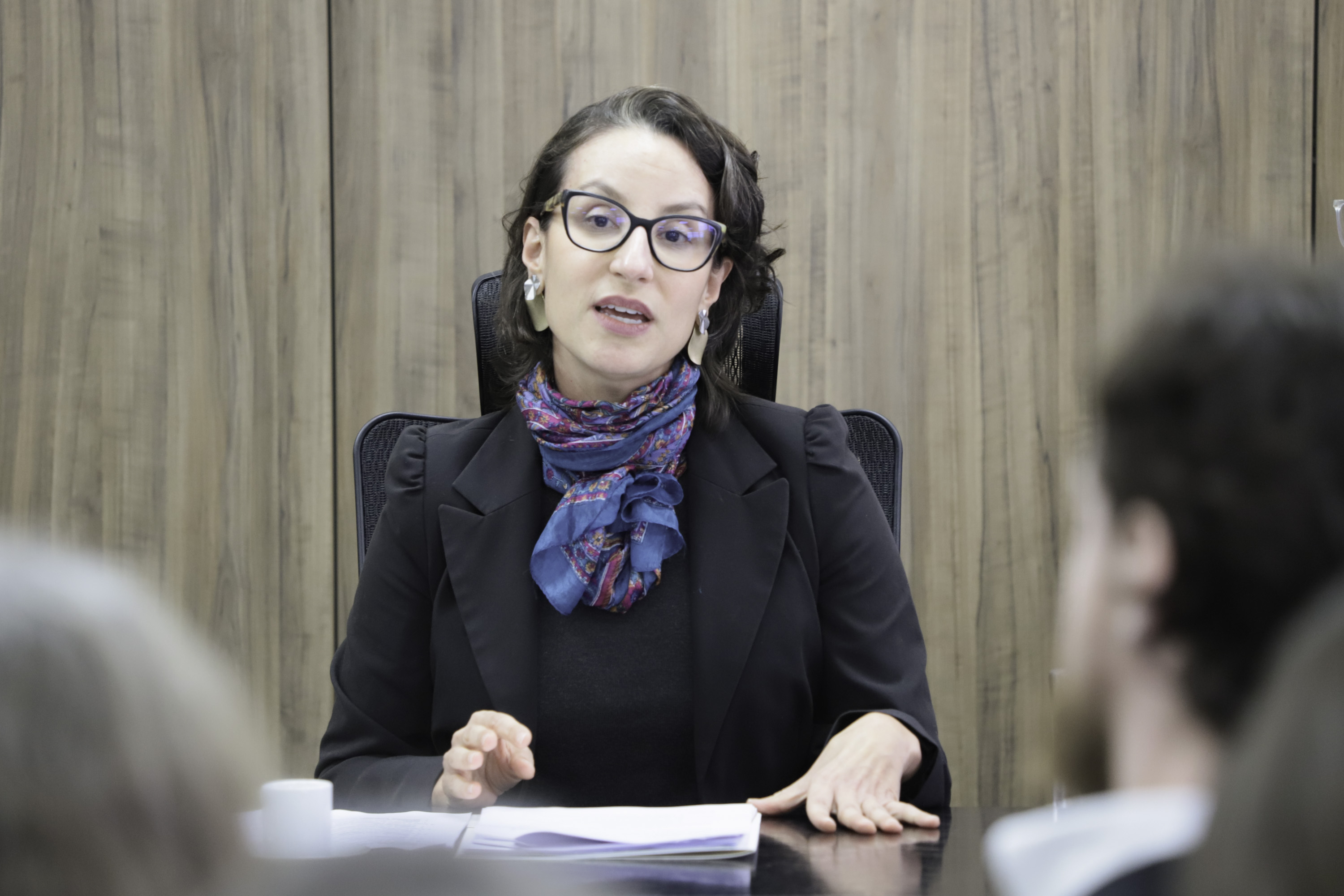 Mariana é branca, fala na mesa oficial com as mãos sobre a mesa. Usa blazer e camisa preta e um cachecol colorido. Tem cabelo curto escuro. 