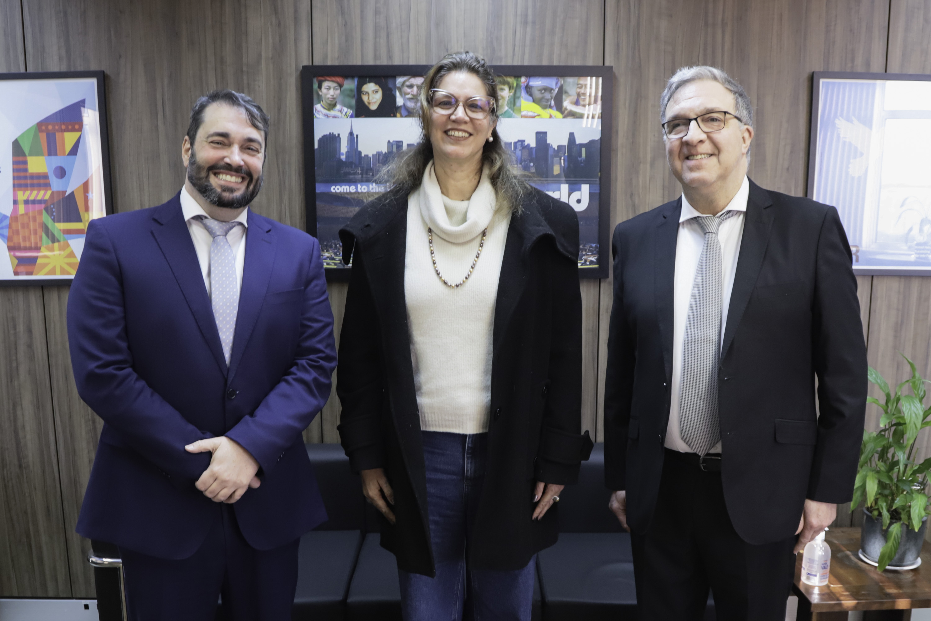 Dois magistados e a magistrada posam sorrindo para a foto, ela aparece ao centro.
