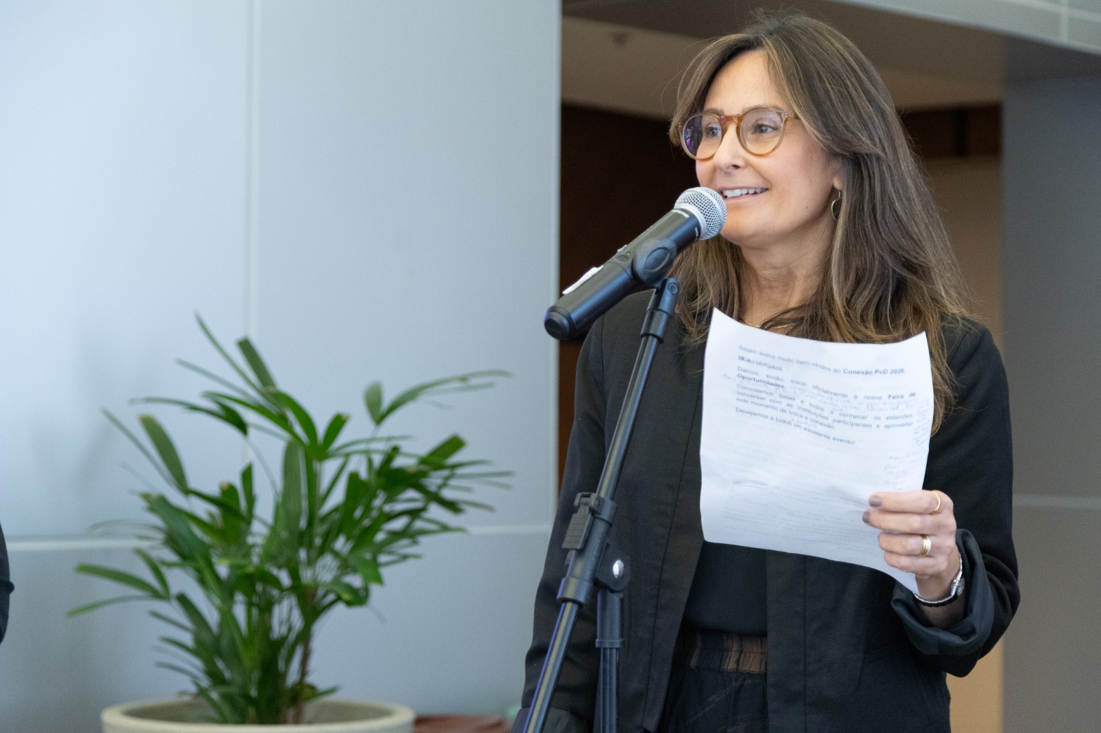 Luciana fala ao microfone sorrindo, segurando uma folha na mão esquerda