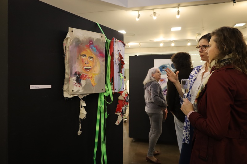 Imagem de duas mulheres observando uma das obras da exposição. As duas mulheres estão de pé, uma delas gesticula com a mão, fazendo um movimento leve de expansão.