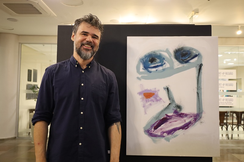 Imagem de João Saikoski ao lado de uma das suas obras. Ele é um homem de pele clara, com cerca de 40 anos, cabelo e barba grisalhos, veste uma camisa azul marinho. Está de pé sorrindo.