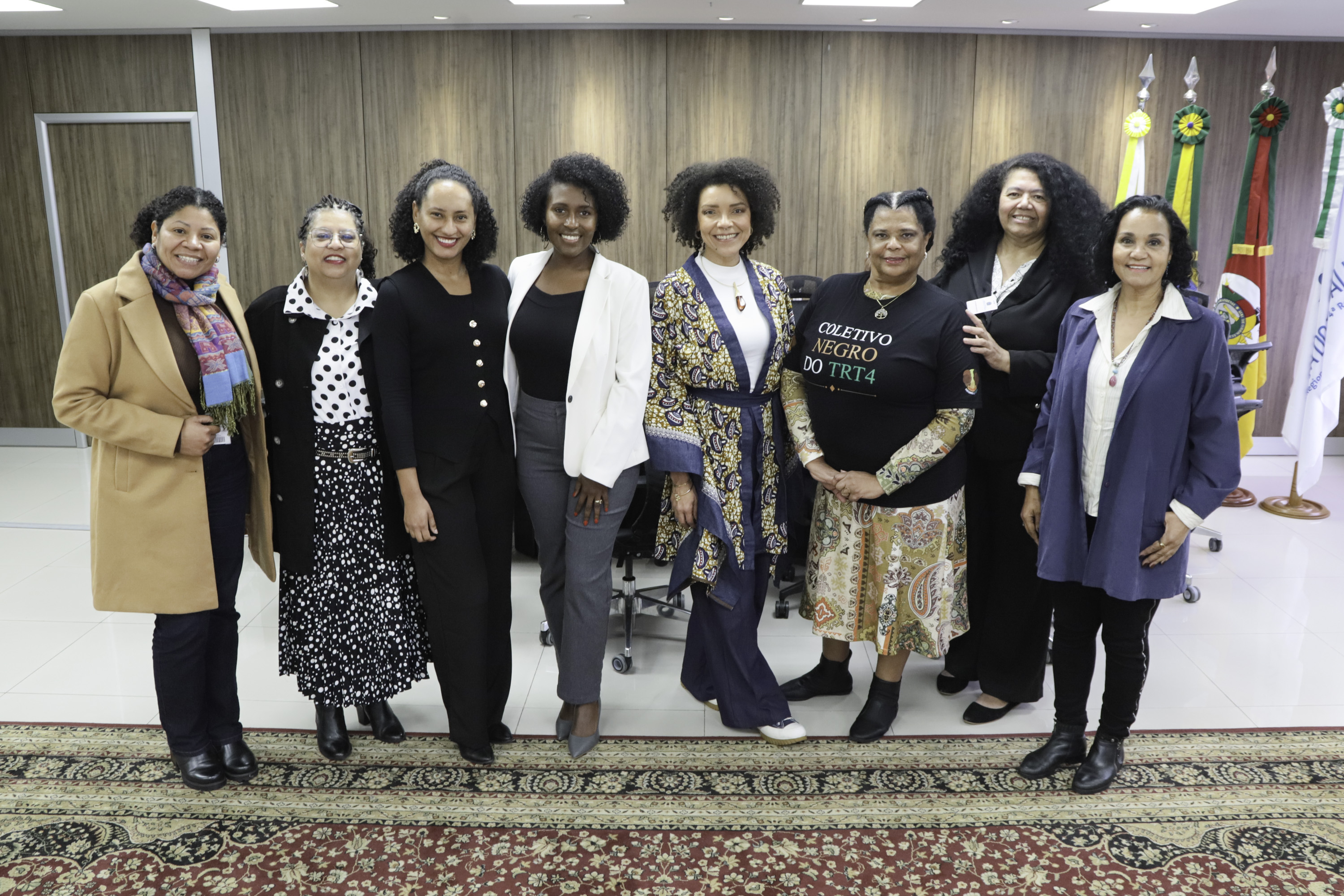 Oito servidoras integrantes do Coletivo posam para a foto sorrindo, de pé, no Salão Nobre da Presidência do Tribunal. Ao fundo aparecem quatro bandeiras.