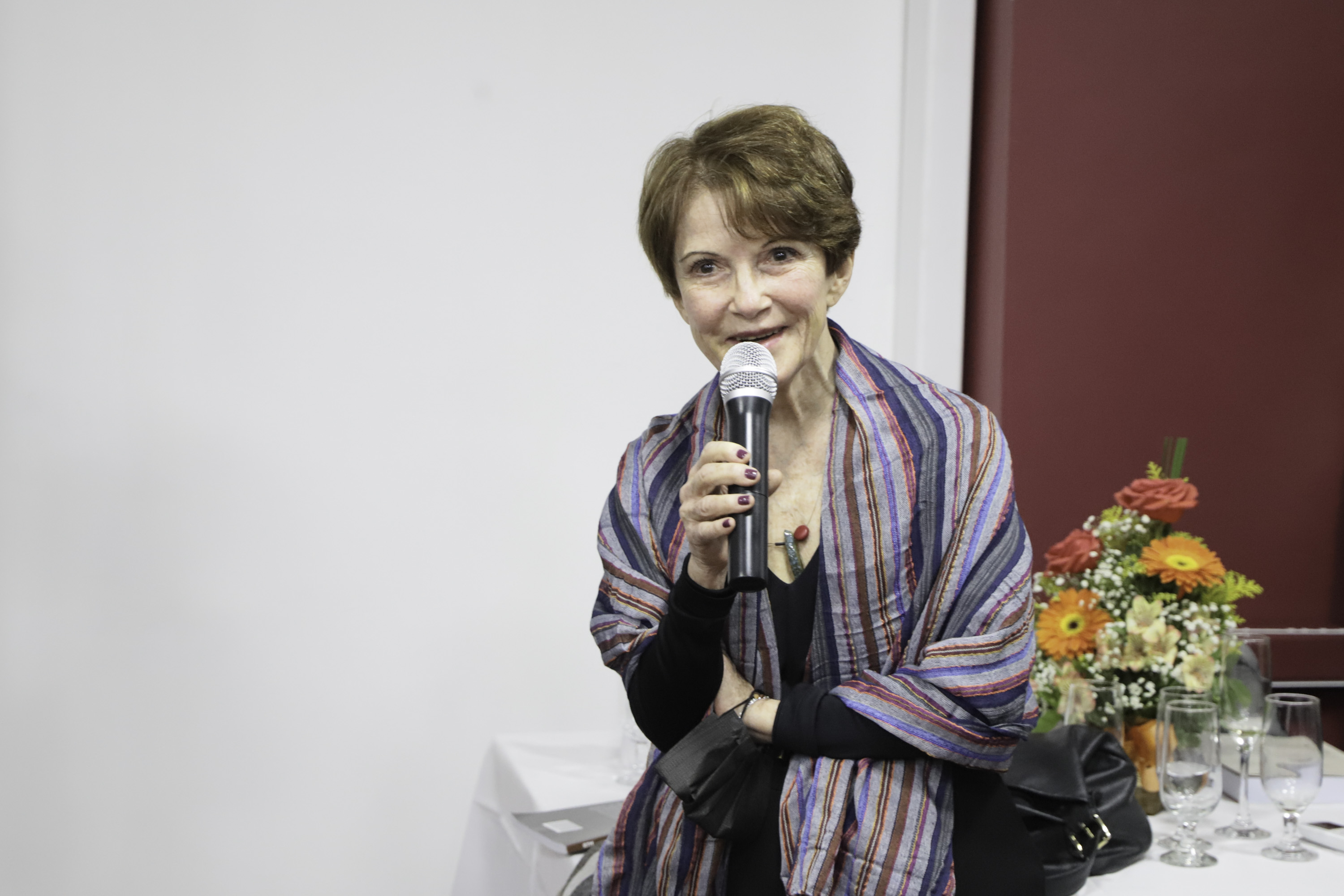Magda fala ao microfone. Atrás dela há uma mesa com um arranjo de flores.