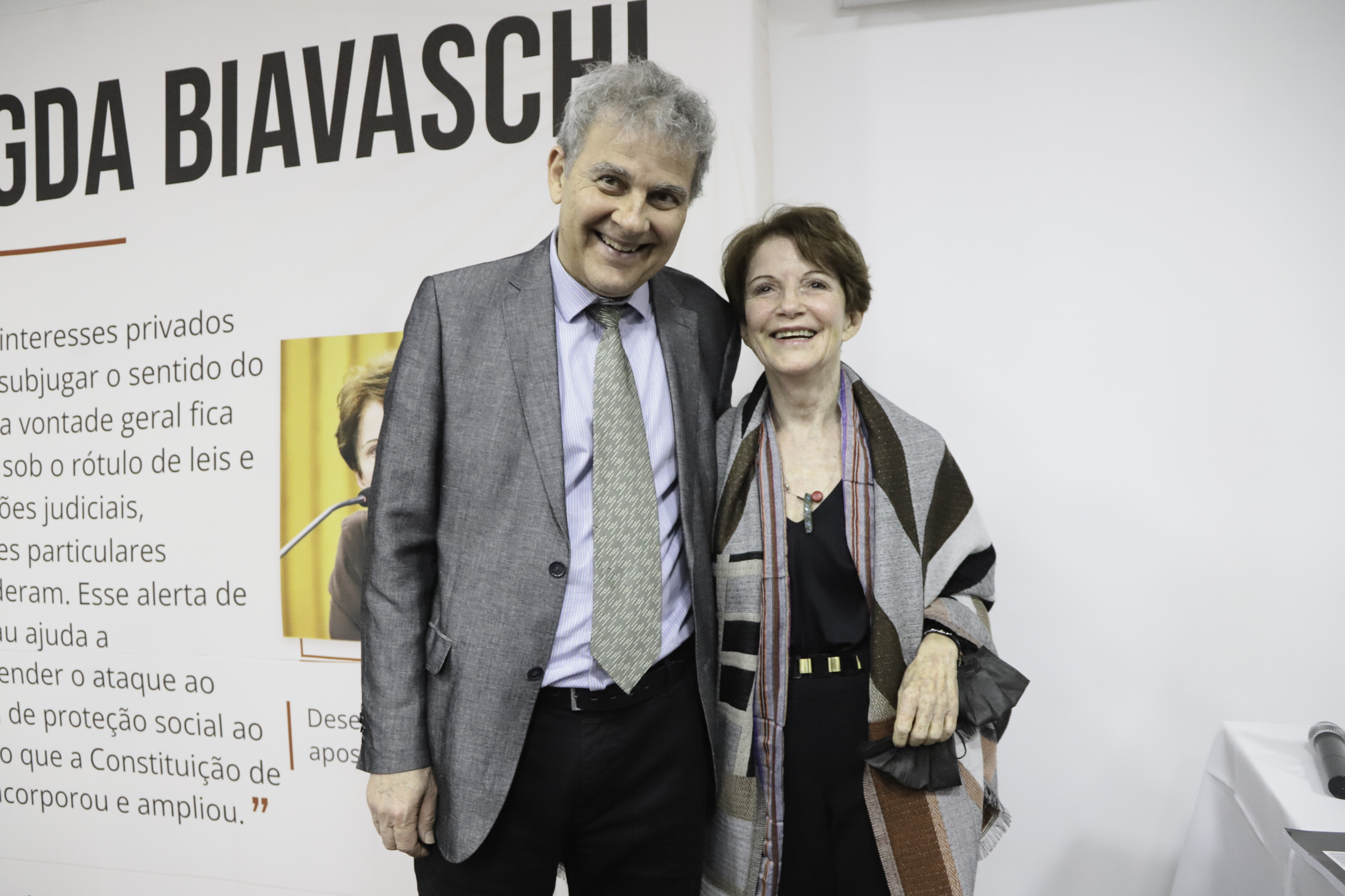 Fraga e Magda sorriem para a foto, atrás aparece um painel com a arte do evento.