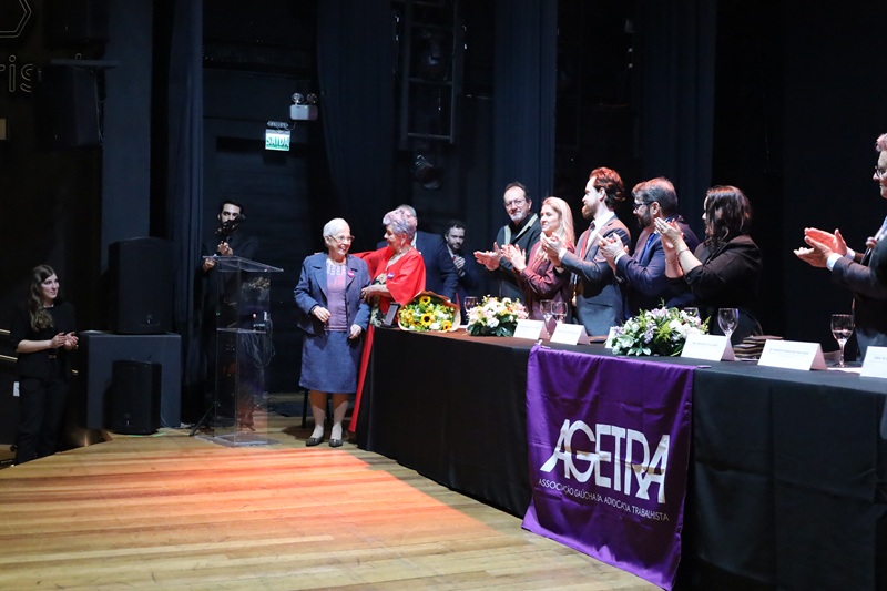 A foto mostra a juíza aposentada e advogada Antônia Mara Vieira Loguercio sendo homenageada no palco, e aplaudida pelos componentes da mesa de abertura.