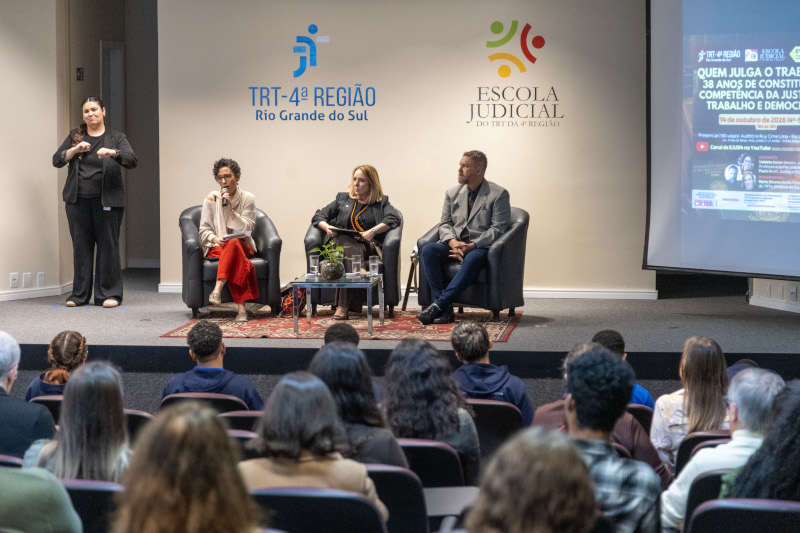 A fotografia mostra todos os participantes sentados no palco e a plateia, de costas.