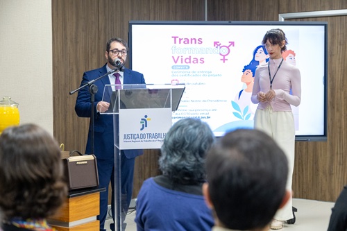 Imagem de Alexandre falando ao microfone, ao lado de Sarah. Ele é um homem de meia-idade, de pele clara, cabelos e barba escuros, veste um terno azul, camisa branca e gravata vermelha, também usa óculos.