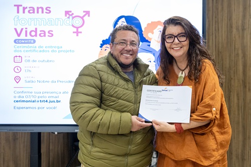 Imagem da juiza Lucia segurando o certificado junto com o formando Matheus. Eles estão de pé, sorrindo. Lucia é uma mulher de 56 anos, cabelo escuro longo, veste uma blusa laranja e usa óculos. Matheus é um homem que aparenta ter cerca de 30 anos, usa óculos e uma jaqueta caqui.