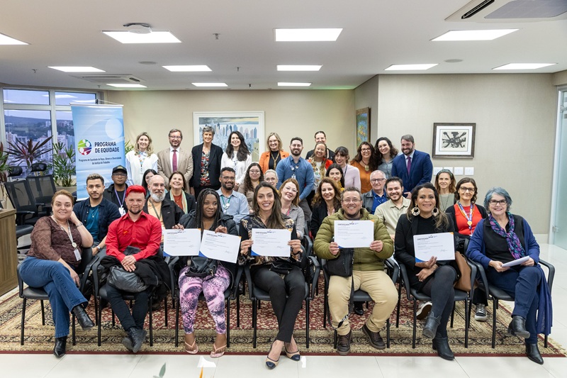 Imagem de todos os presentes na cerimônia reunidos. Muitos estão sentados e na fileira de trás estão de pé alguns dos representantes do TRT-RS, dentre eles, o presidente. Na fileira de cadeiras da frente estão os quatro formandos exibindo seus certificados.