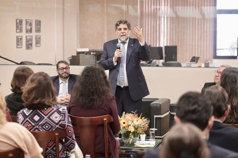 A foto mostra o ministro Caputo em pé, palestrando, ao lado do presidente Alexandre e do desembargador Cassou.
