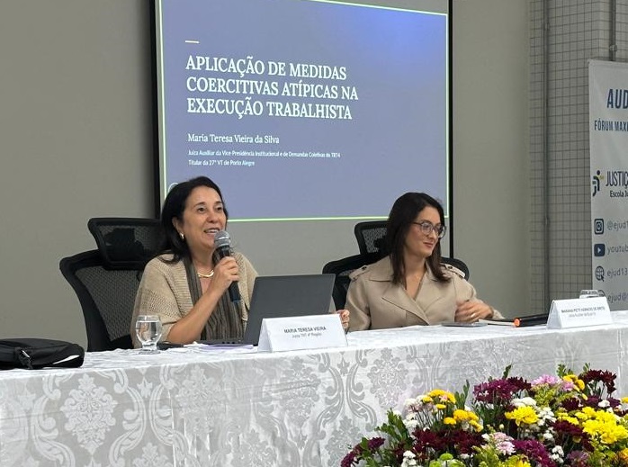 Juíza Maria Teresa fala ao microfone sentada à mesa oficial do evento.