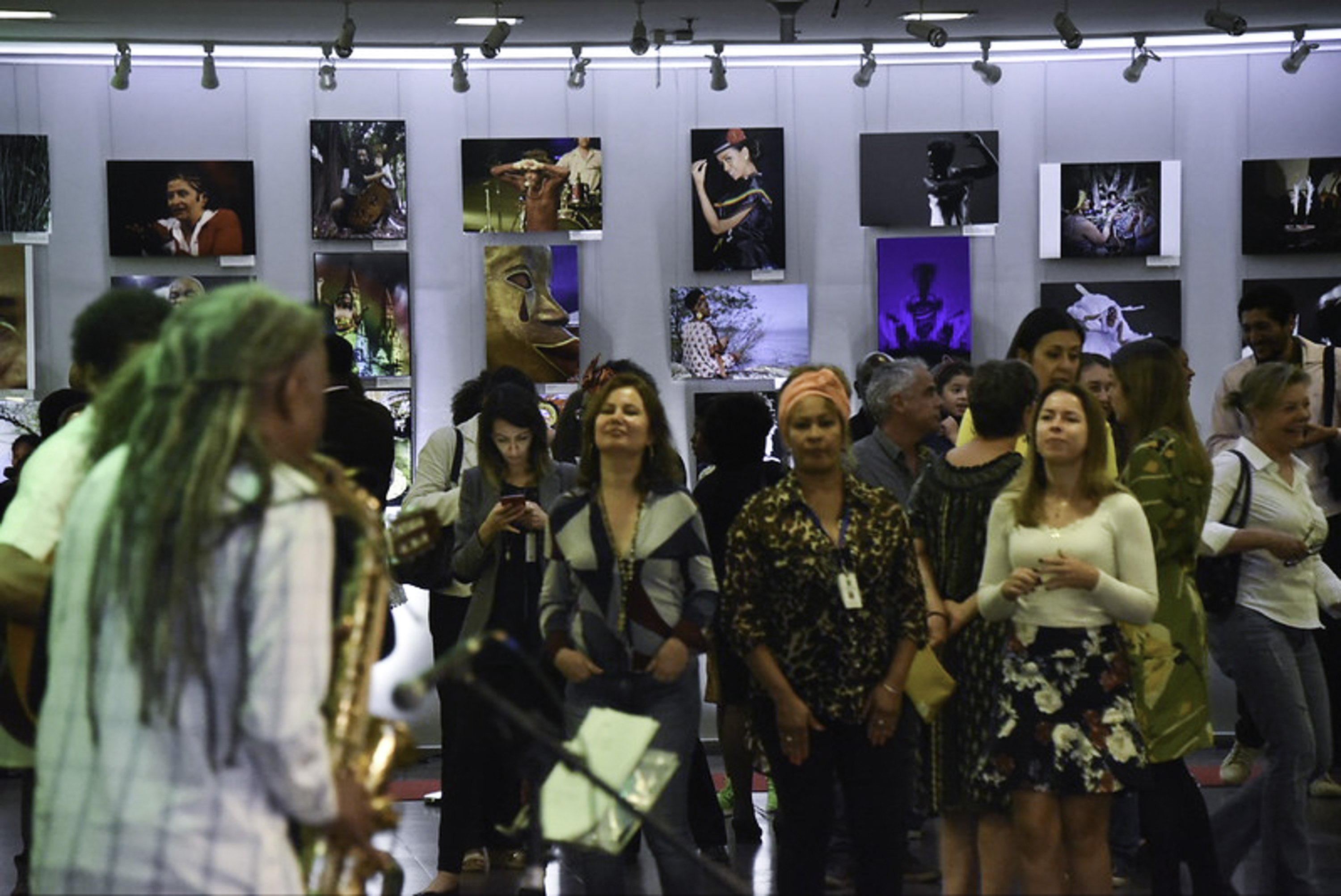 Músico toca saxofone na esquerda, público na direita e fotos em exposição ao fundo.