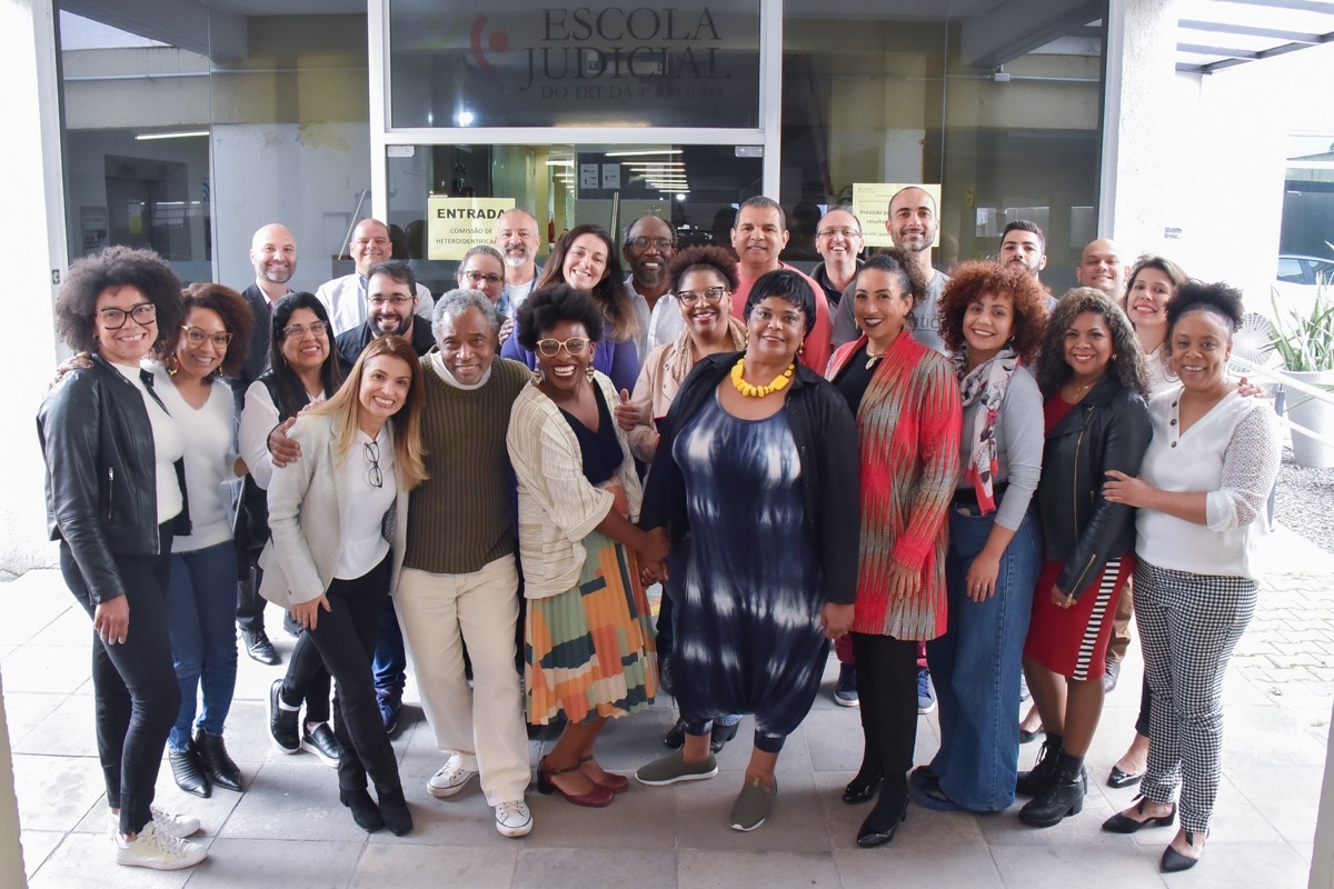 25 pessoas posam para a foto de pé e sorrindo em frente ao prédio da Escola Judicia..