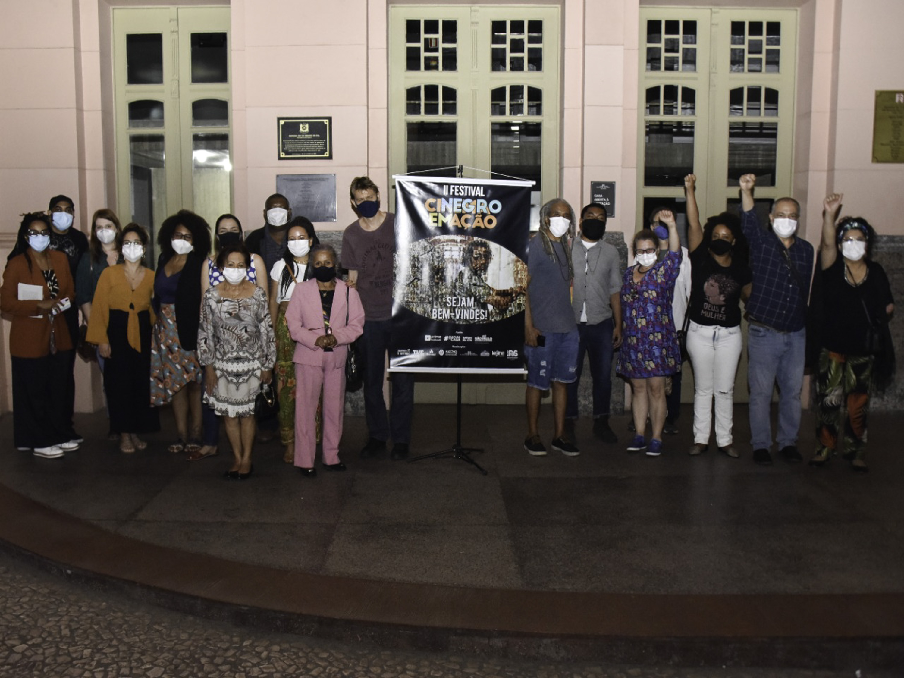 Diversasa pessoas posam usando máscara com o cartaz do festival ao centro. Ao fundo aparece o prédio rosa da Casa de Cultura.
