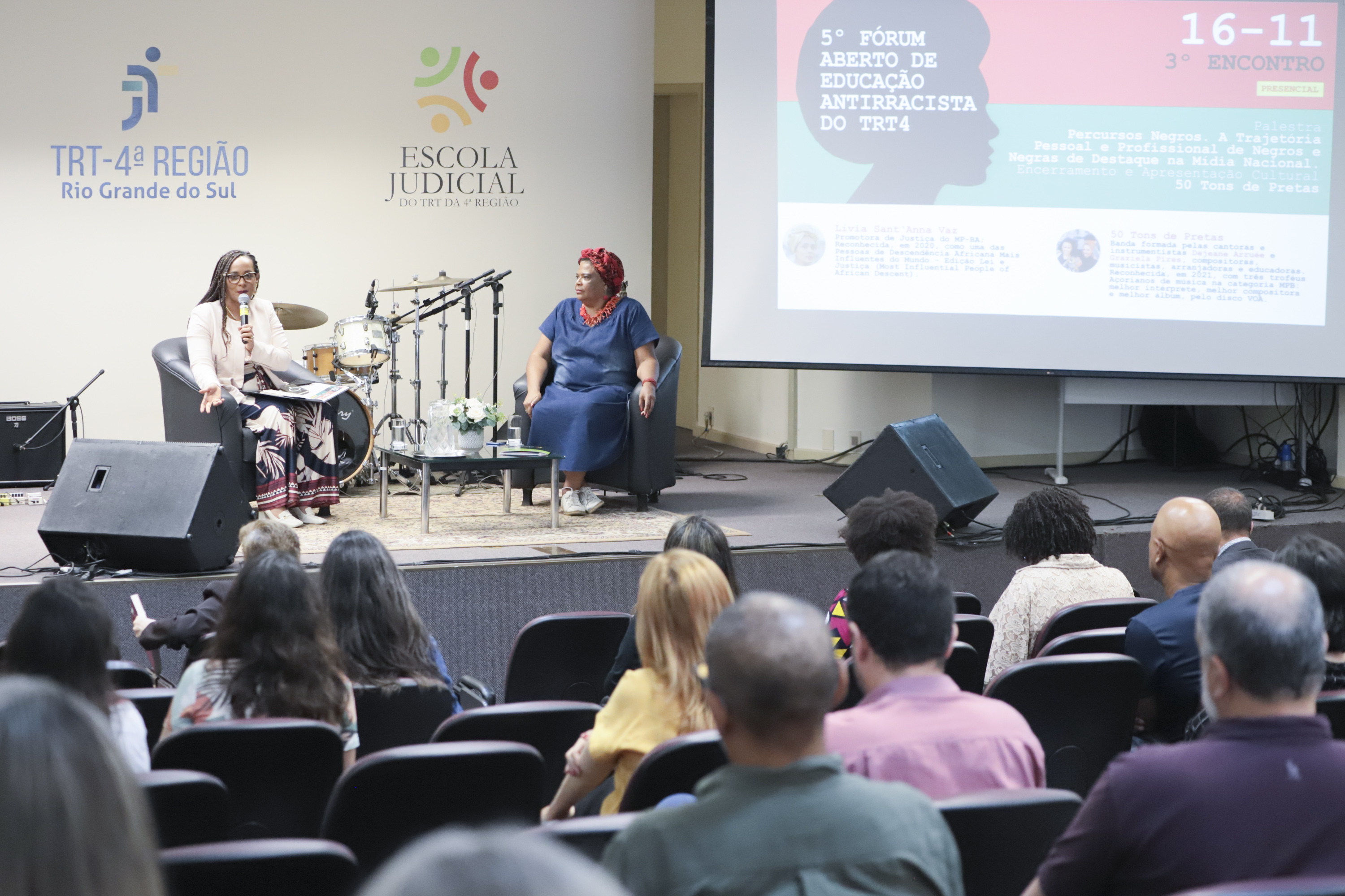 Duas pessoas aparecem no palco ao fundo. Na direita aparece o logo do evento. Em primeiro plano aparece o público de costas