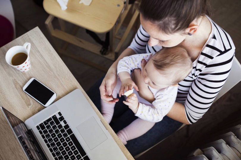 Imagem superior de uma jovem mãe trabalhando com o filho bebê no colo. Ambos têm a pele branca e cabelos castanhos. Em frente aos dois, há uma mesa com notebook e uma xícara de café.