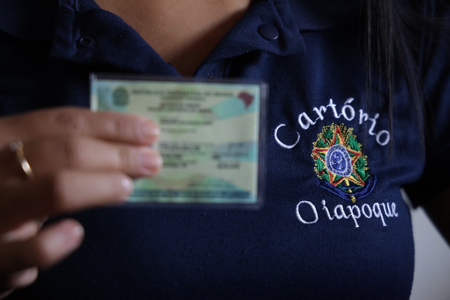 Uma pessoa segura em primeiro plano um documento de identidade brasileiro plastificado, que se encontra desfocado. Em foco, veste um polo azul-escuro com o brasão da República Federativa do Brasil bordado e as palavras «Cartório» acima e «Oiapoque» abaixo. Na mão que segura o documento, destaca-se um anel simples no dedo.