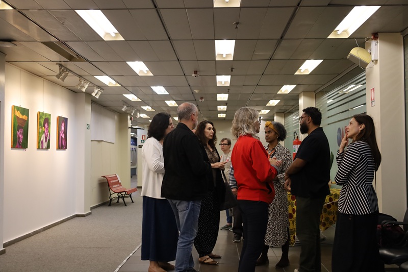 Imagem geral da abertura da exposição. A imagem mostra um grupo de pessoas conversando reunidas à mesa do coquetel à direita. E a exposição de quadros à esquerda.