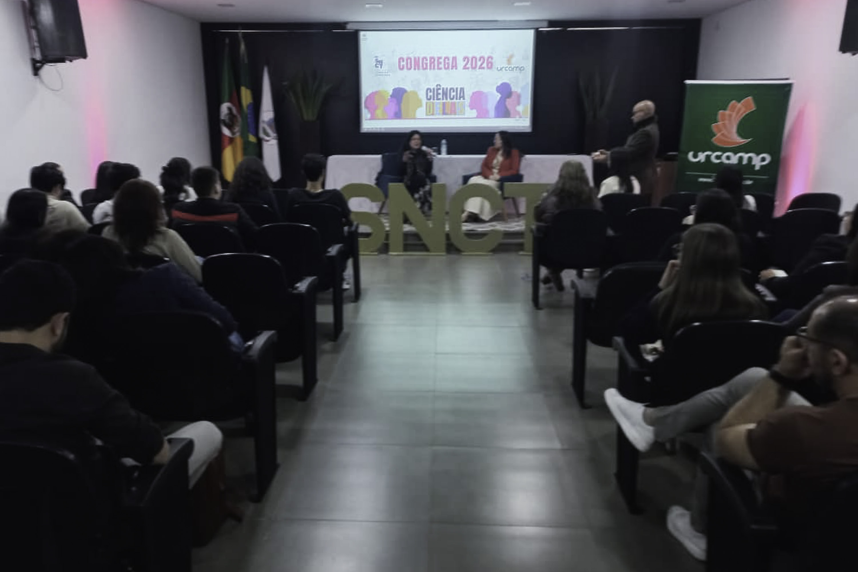 Palestraantes ao fundo, com público de costas nas laterais da imagem. 