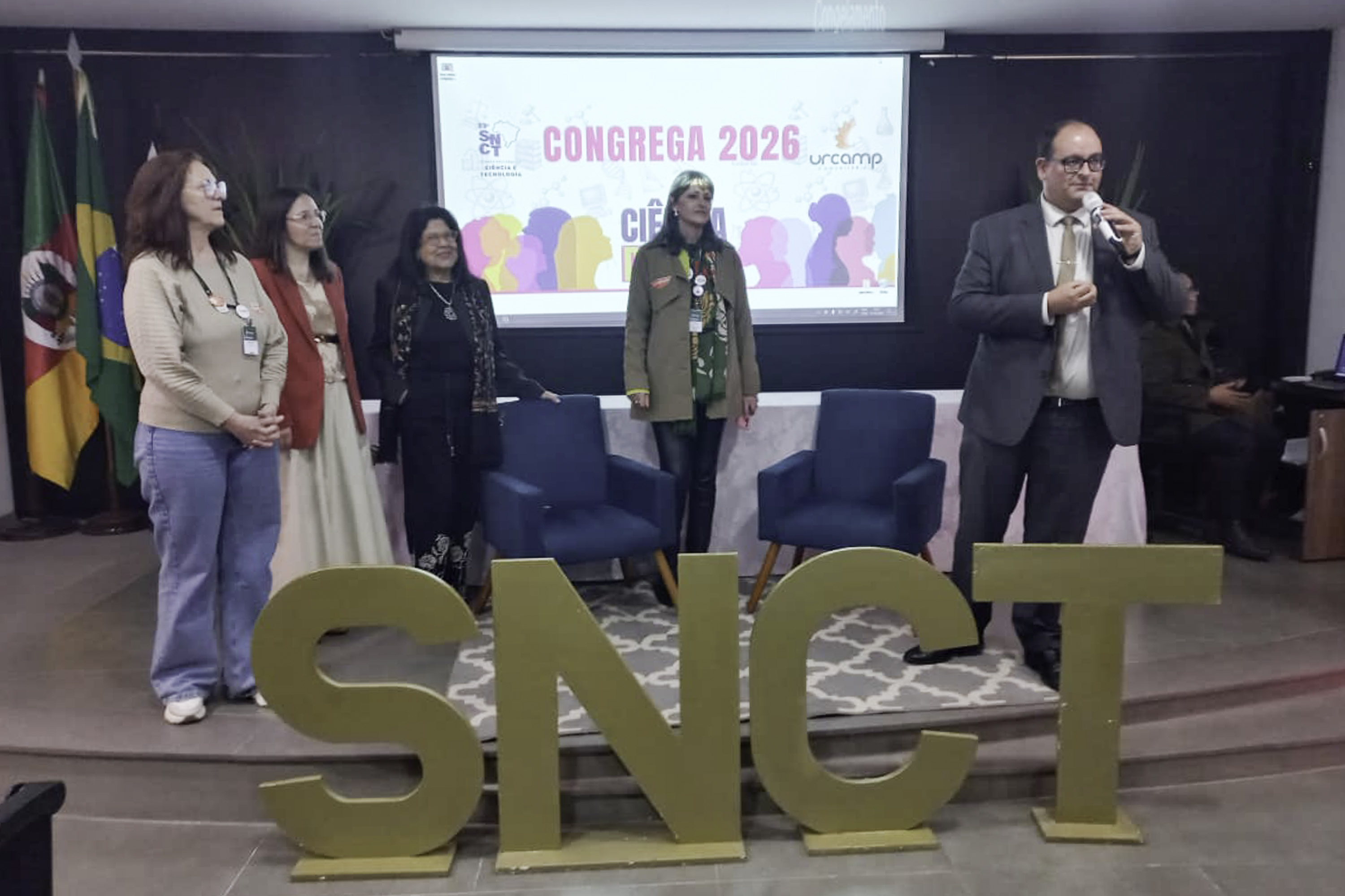 Cinco pessoas aparecem no palco, com o reitor no canto direito falando ao microfone. 