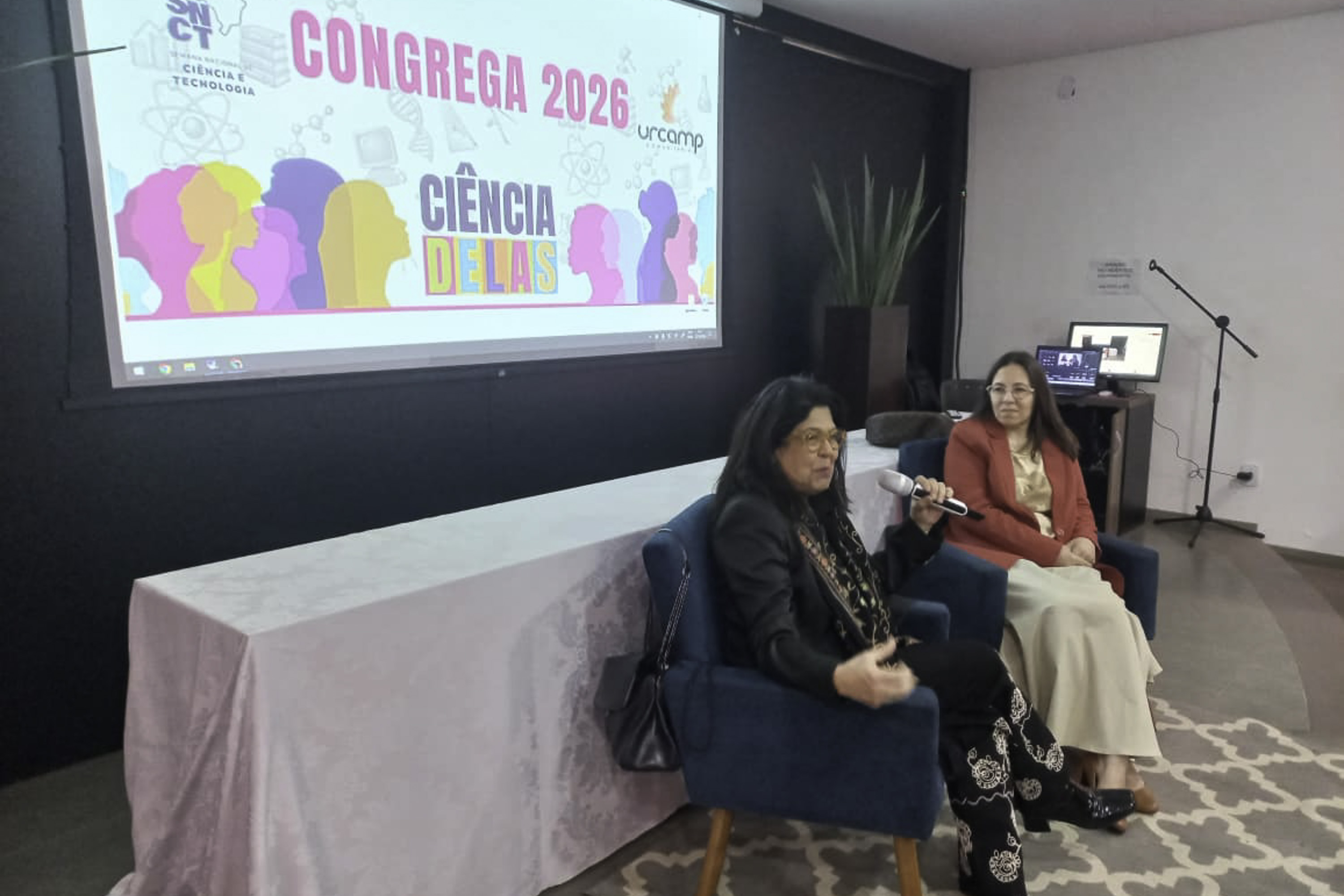 Brígida fala em primeiro plano com Marcele a seu lado. Atrás delas aparce o telão com a arte do evento