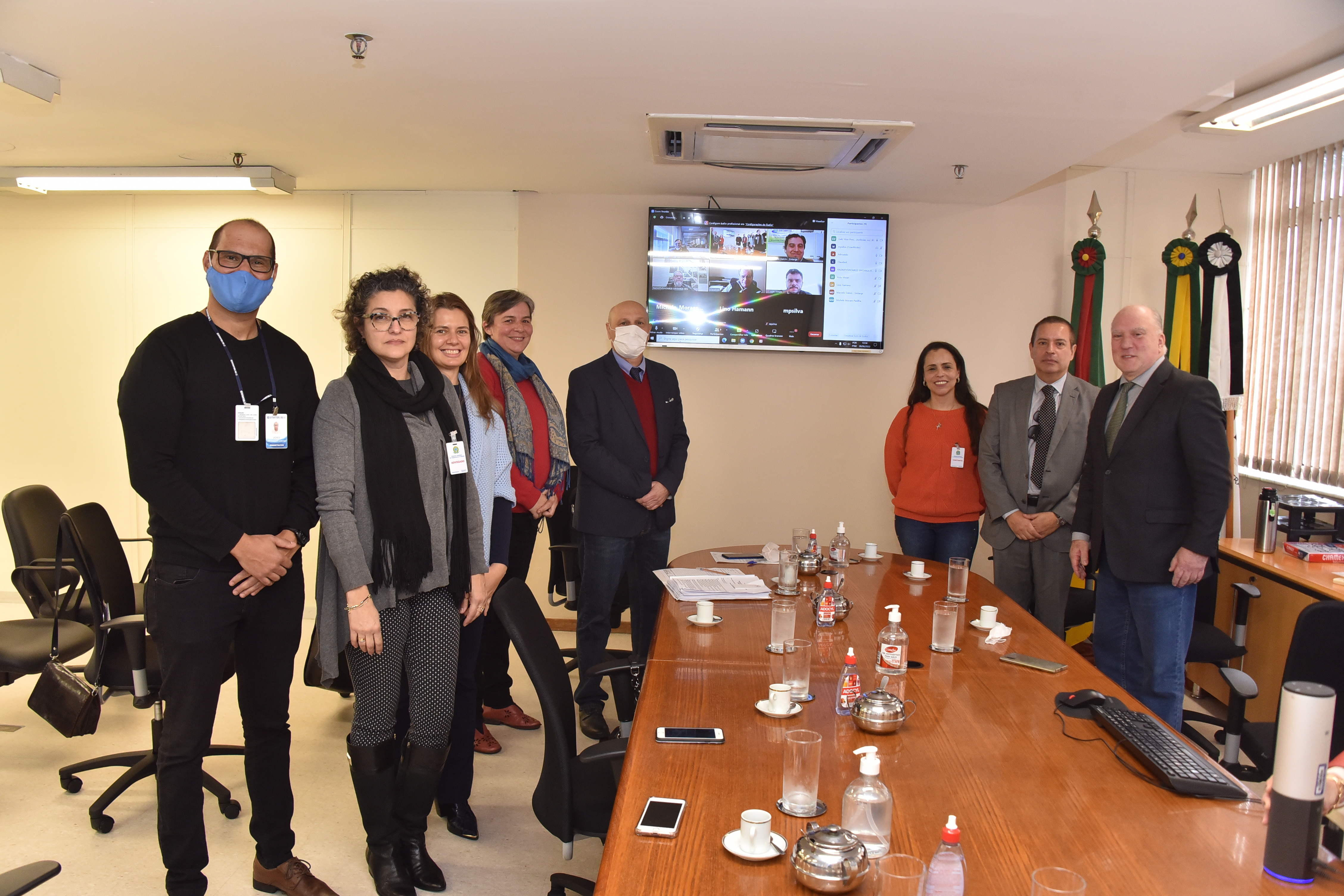 Foto dos participantes em pé, posando para a foto. Estão perfilados ao lado da mesa, e ao fundo aparece a tela com os participantes por videoconferência.