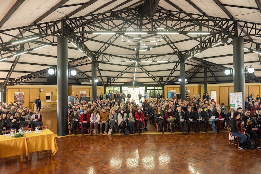 Foto  do público no evento, sentados nas cadeiras em um amplo salão. No canto direito aparece a orquestra de jovens artistas. 