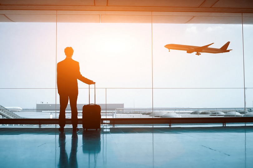 Foto ilustrativa de um homem com mala na mão dentro de aeroporto. Ao fundo, no céu, passa uma aeronave.