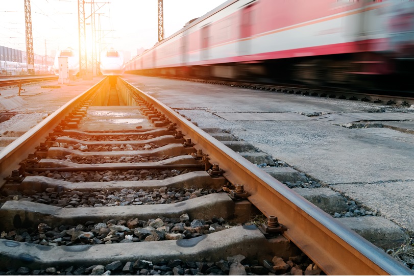 Foto ilustrativa mostra trilhos de trem em primeiro plano. Ao lado, há um trem passando rapidamente. A cor é vermelha e a imagem está desfocada.