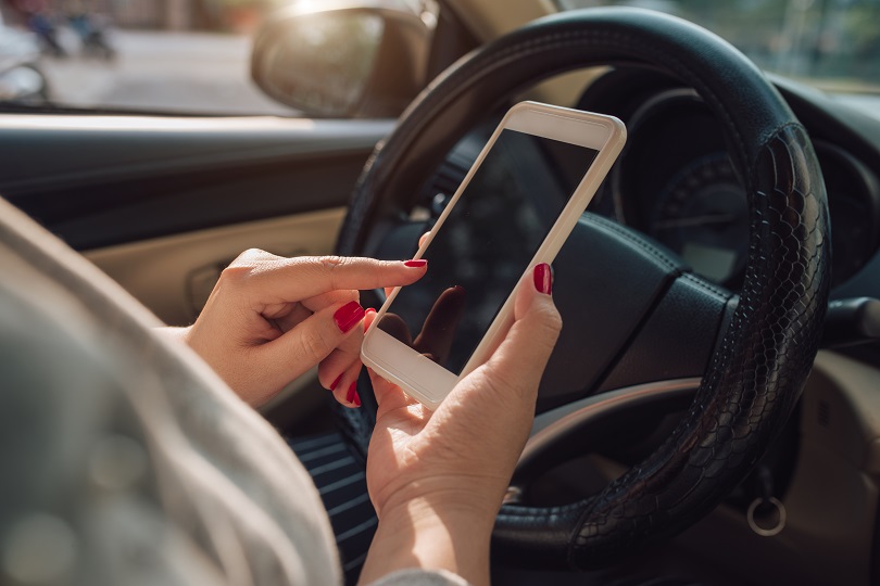 Foto ilustrativa mostra mão de mulher usando smartphone. A tela do celular está escura. Ela usa uma blusa bege e tem as unhas pintadas de vermelho. Está dentro de um carro e a imagem mostra a direção, na cor preta. 