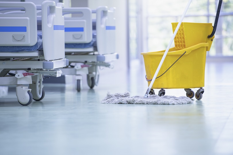 Foto ilustrativa mostra detalhe de camas de hospital, carrinho de hospital e rodo de limpeza. As camas são nas cores cinza claro e azul e o carrinho é amarelo.