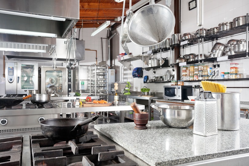 Foto ilustrativa mostra cozinha comercial. Há fogões industriais, panelas e demais utensílios de cozinha dispostos em prateleiras e suportes aéreos. Predominam as cores cinza e prata. 