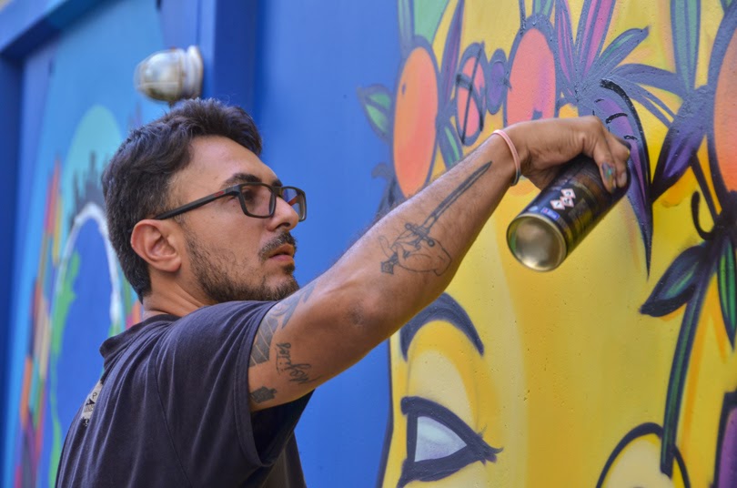 JotaPê Pax em close trabalhando no mural. Ele segura um spray e aplica a tina sobre o muro