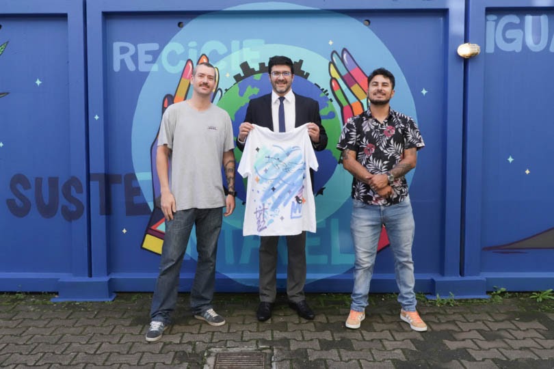 Os dois artistas posam para a foto em frente ao mural com o presidente Rossal entre eles. O presidente segura uma camiseta presenteada pelos artistas