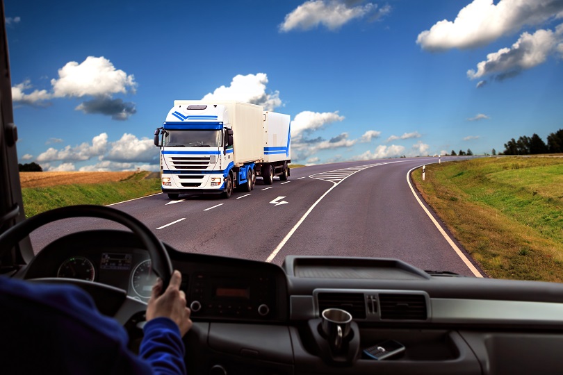 Foto mostra vista da estrada, a partir de cockpit de caminhão. Pode-se ver os braços do condutor do veículo e o painel na cor preta. Há um céu nublado e outro caminhão, nas cores azul e branco, em trânsito.