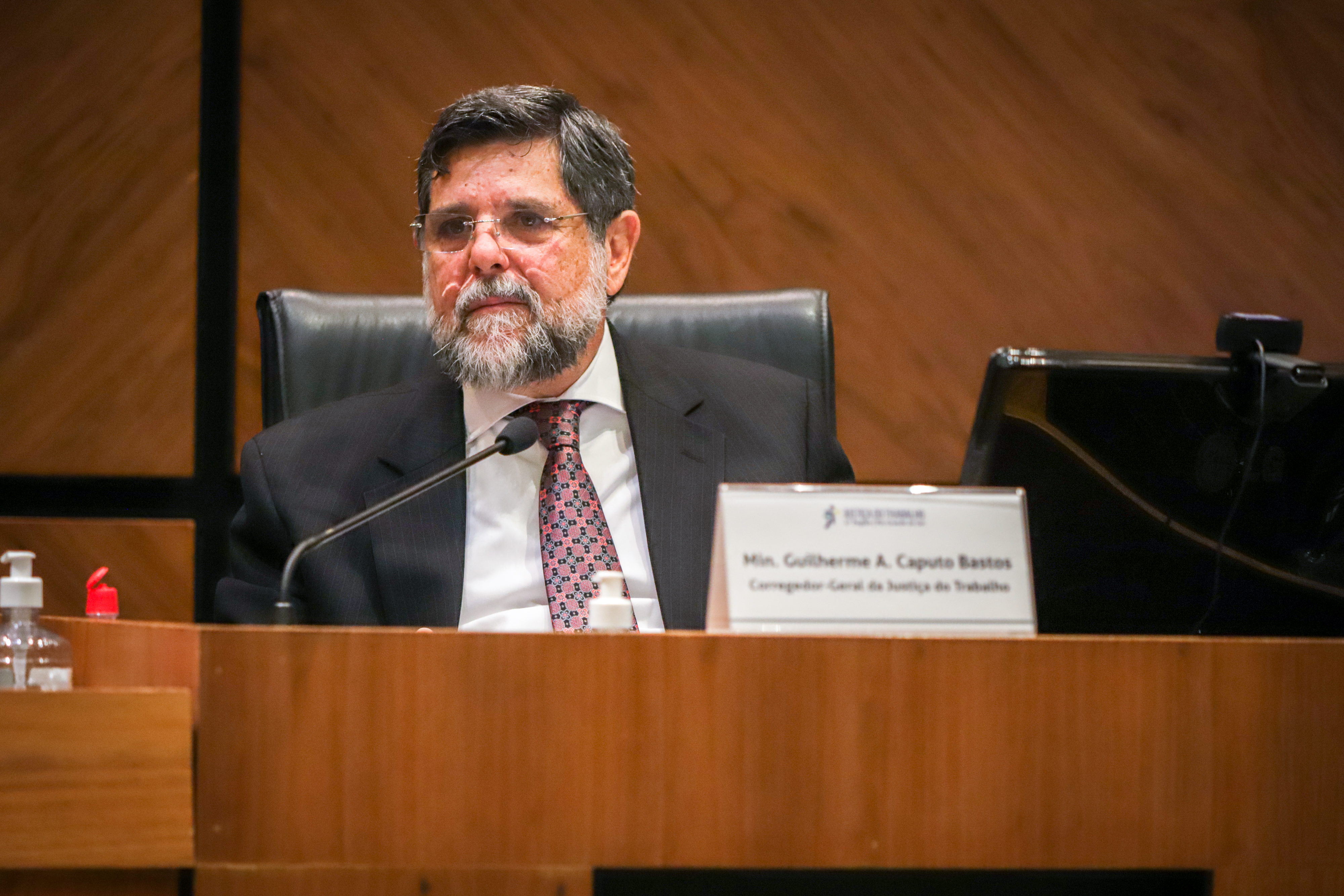 ministro Caputo Bastos é banco, de barba grisalha e cabelo escuro. usa óculos e aparece atrás do microfone. 